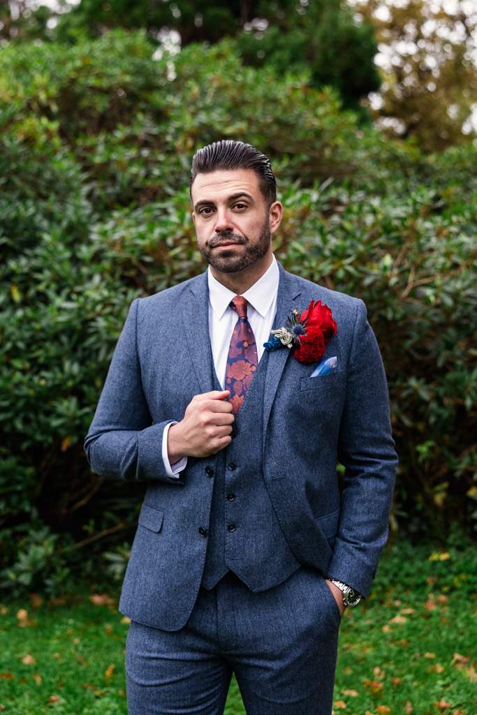Portrait of groom in a blue suit with a red rose boutonniere standing outdoors in front of green bushes on the grounds of Markree Castle