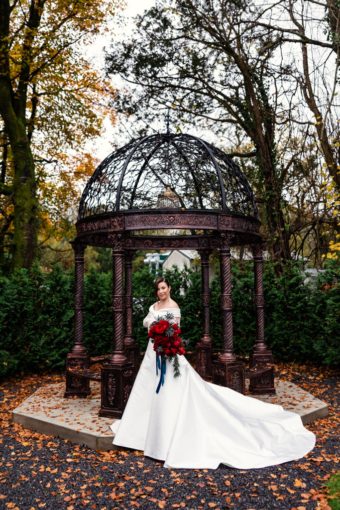 Portrait of bride in a white gown holding red bouquet standing under a gazebo, surrounded by autumn trees and leaves at Markree Castle