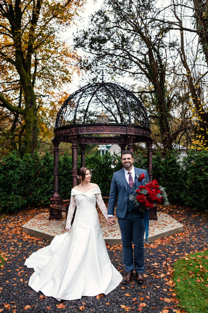 Bride and groom stand holding hands in front of a decorative gazebo surrounded by autumn trees outside Markree Castle