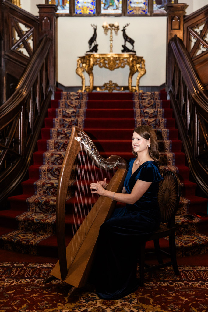 Woman in a blue dress playing a harp on a grand staircase with red carpet and stained glass windows inside Markree Castle