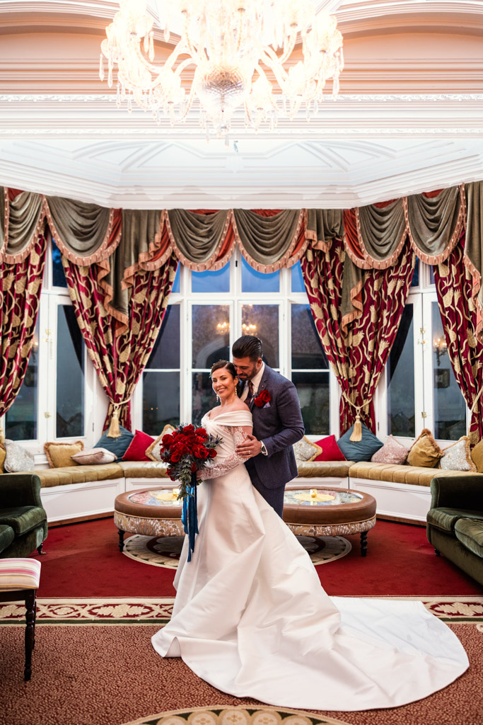 A bride and groom stand smiling in an elegant, ornate room with patterned curtains and a chandelier inside Markree Castle