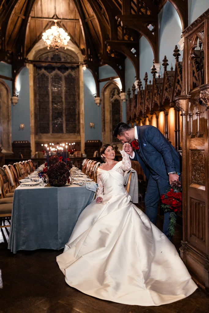 Bride in white gown sits at a decorated table as groom in suit kisses her hand in an ornate hall inside Markree Castle