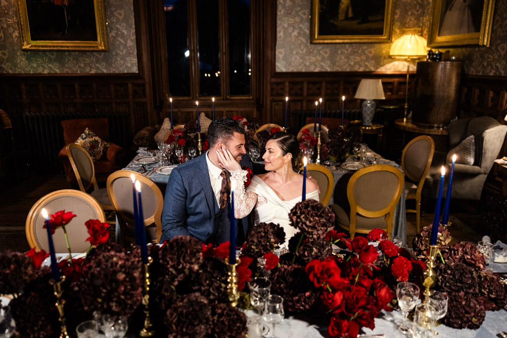 A bride and groom smile at each other, seated at a candlelit table decorated with red flowers inside Markree Castle