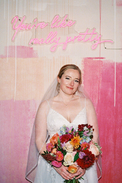 Bride in a white dress holding a colorful bouquet, standing under a neon sign that reads "You're like really pretty" at Lacuna Lofts.