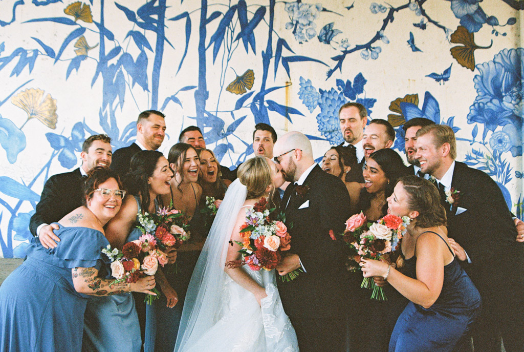 Film photo of bride and groom kissing surrounded by a joyful wedding party in front of a vibrant blue floral mural at Ping Tom Memorial Park