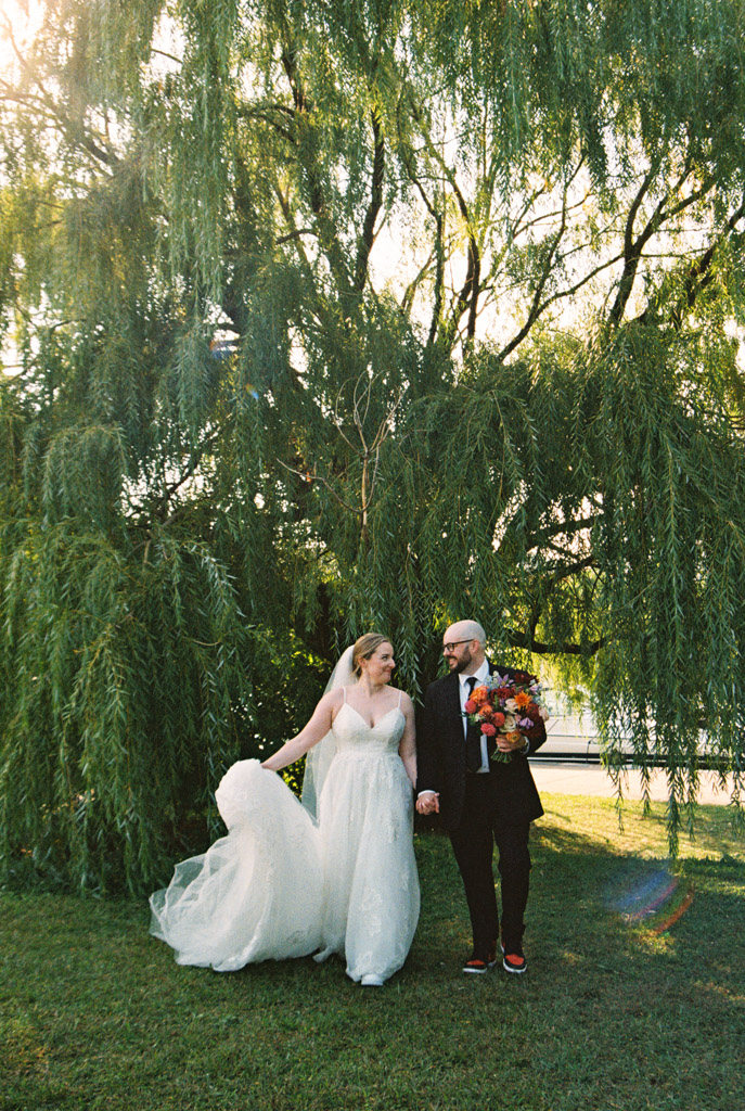 Bride and groom hold hands and smile under a large willow tree at Ping Tom Memorial Park, with sunlight shining through the leaves