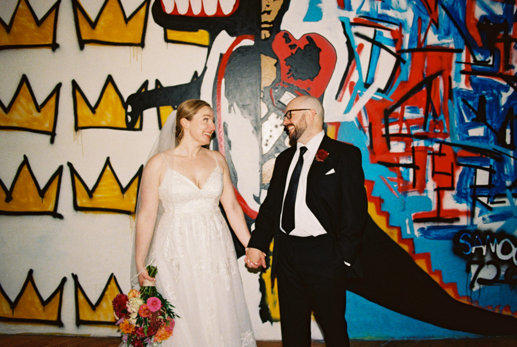 Film photo of bride and groom holding hands, smiling, standing in front of a colorful Basquiat-themed wall mural at Lacuna Lofts