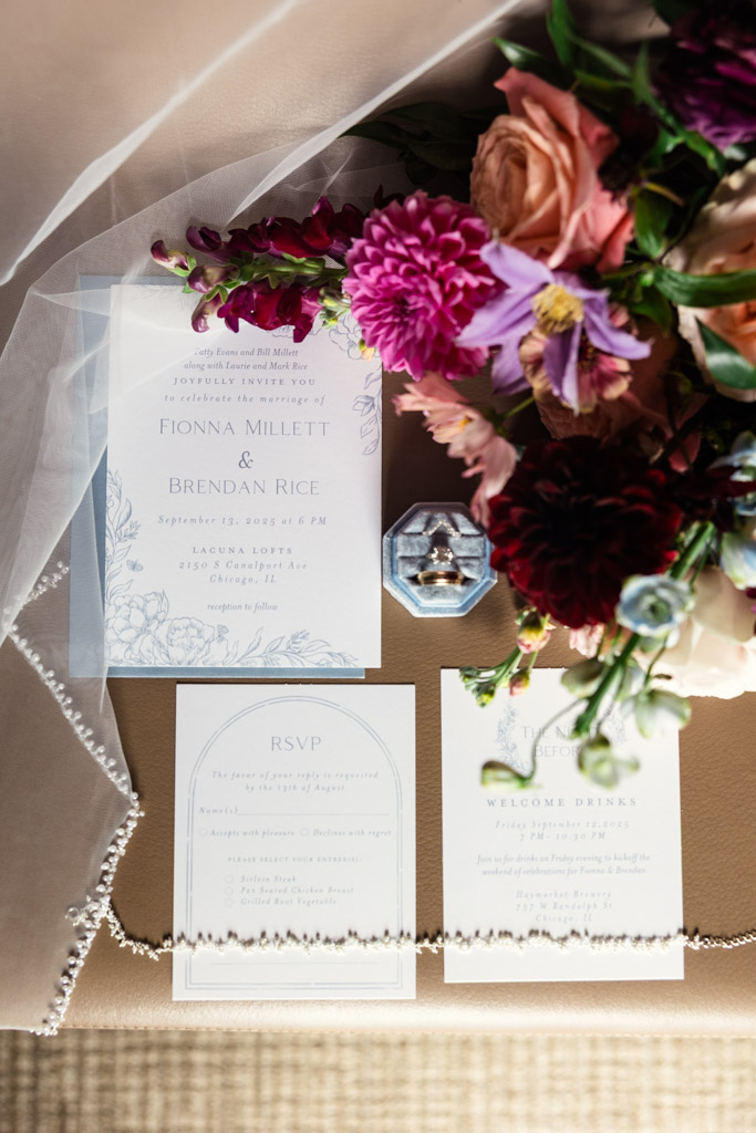 Wedding invitation suite with RSVP card, flowers, rings in a box, and a beaded veil on a beige surface for an wedding celebration at Lacuna Lofts