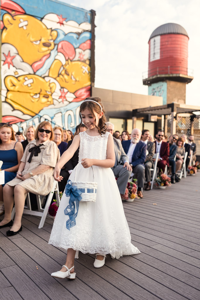 Flower girl in a white dress walks down the aisle of Lacuna Lofts rooftop wedding ceremony space holding a basket, with guests seated on either side