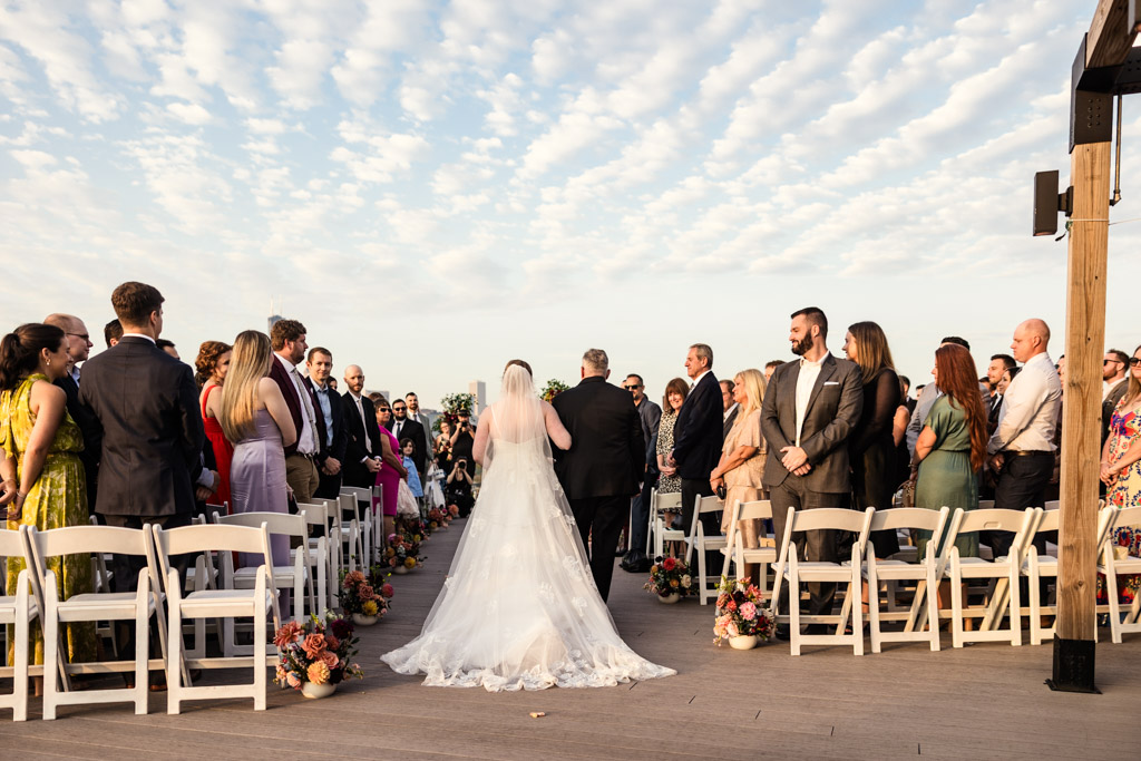 Bride walks down the aisle with her father in the rooftop wedding ceremony space at Lacuna Lofts, guests standing on either side under a blue, lightly clouded sky