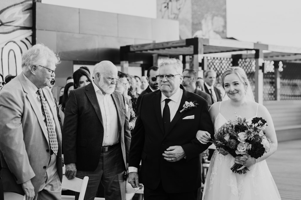 Black and white photo of bride in white dress and her father walking down the aisle, guests watching and smiling during Lacuna Lofts rooftop wedding ceremony
