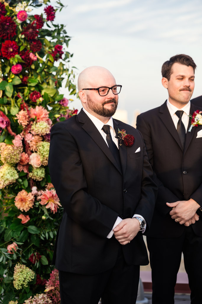 Groom in a black suit watches bride walk down the aisle standing in front of a vibrant floral arrangement during Lacuna Lofts wedding ceremony