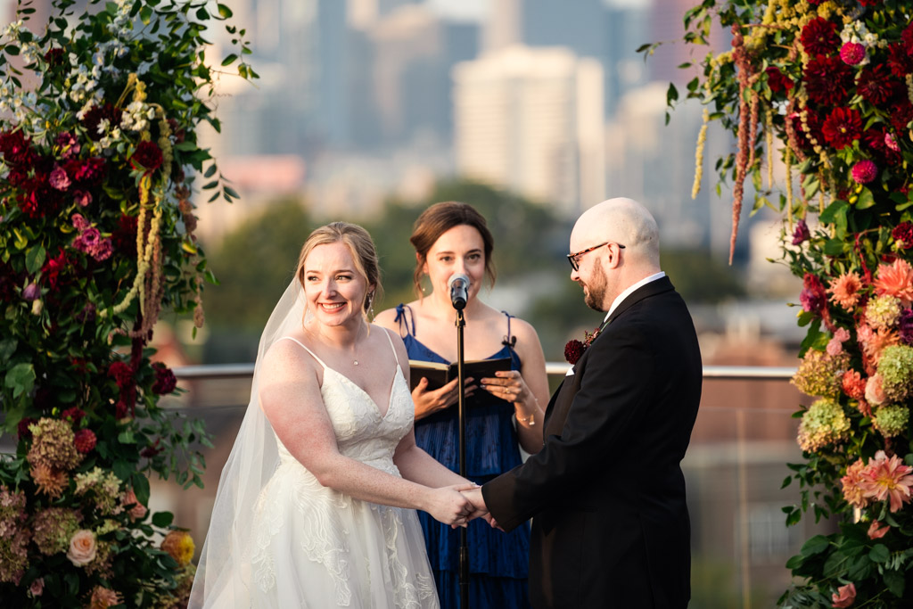 Bride and groom hold hands and smile during an rooftop wedding ceremony at Lacuna Lofts while the officiant speaks, flanked by floral arches