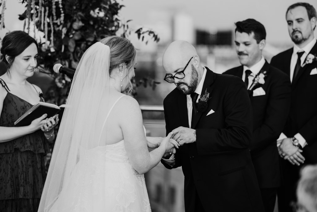 Black and white photo of groom placing ring on the bride’s finger during rooftop wedding ceremony at Lacuna Lofts