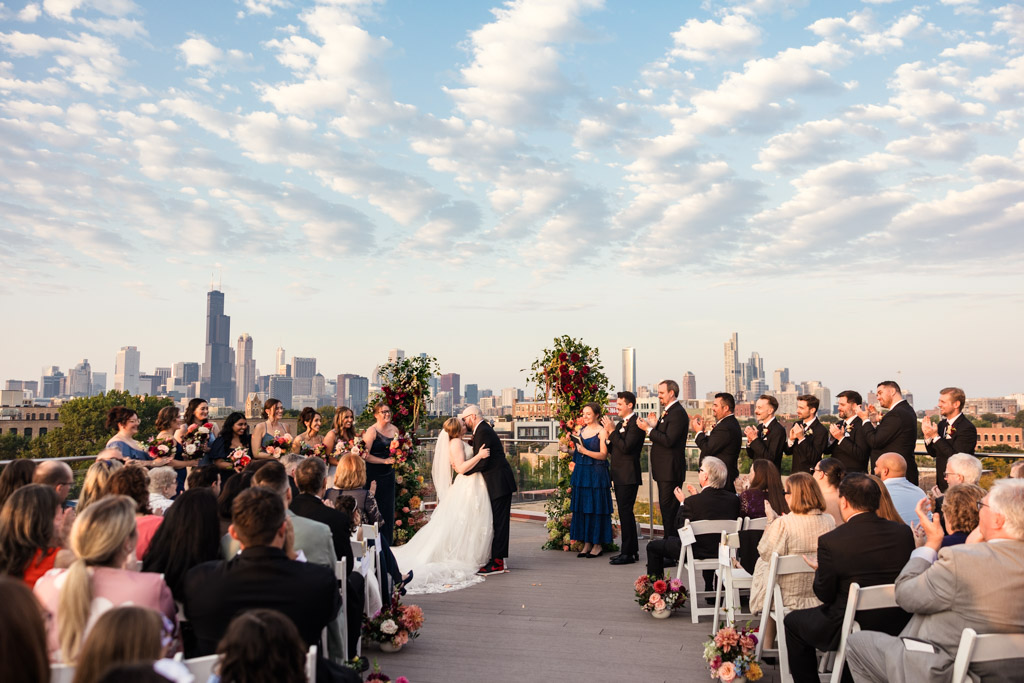 A couple kisses during their rooftop wedding ceremony at Lacuna Lofts, while guests applaud with the Chicago skyline in the background
