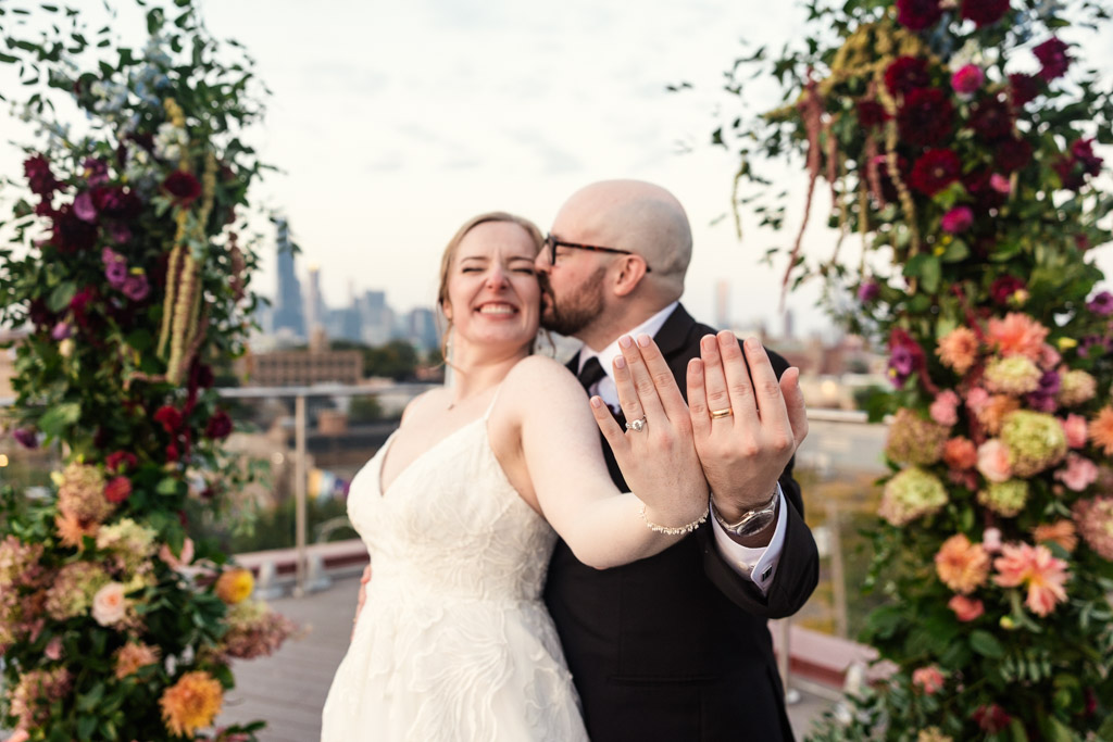 Bride smiles and shows off her wedding ring as the groom kisses her cheek after their rooftop wedding ceremony at Lacuna Lofts