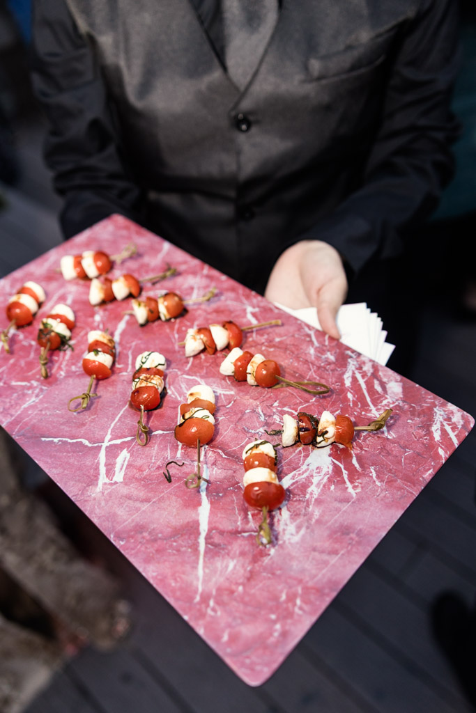 A person in a dark vest holds a pink tray with skewered appetizers of tomato, mozzarella, and basil for wedding reception at Lacuna Lofts