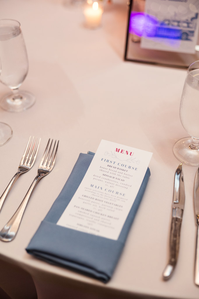 Wedding reception guest place setting with dinner menu on a blue napkin, silverware and water glasses on a white tablecloth at Lacuna Lofts
