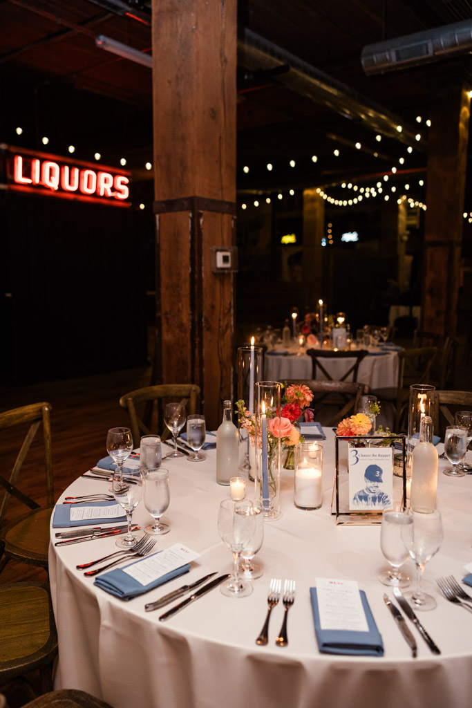 Guest table set with candles, flowers, and menus for Lacuna Lofts wedding reception, a neon sign glows in the background