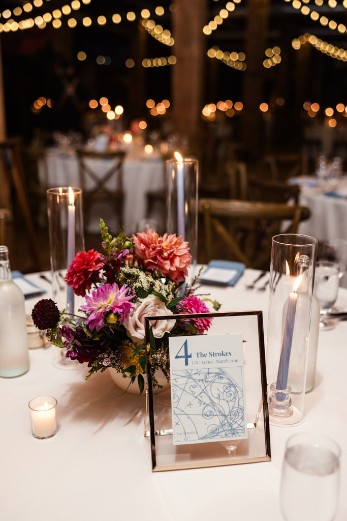 Guest table with flowers, candles, and a framed custom concert-themed table sign under string lights at night for wedding reception at Lacuna Lofts