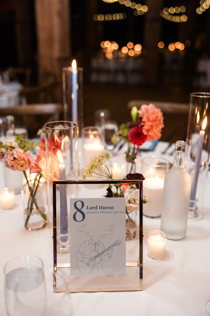 Guest table decorated with candles, flowers, and a framed custom concert-themed table sign for Lacuna Lofts wedding reception