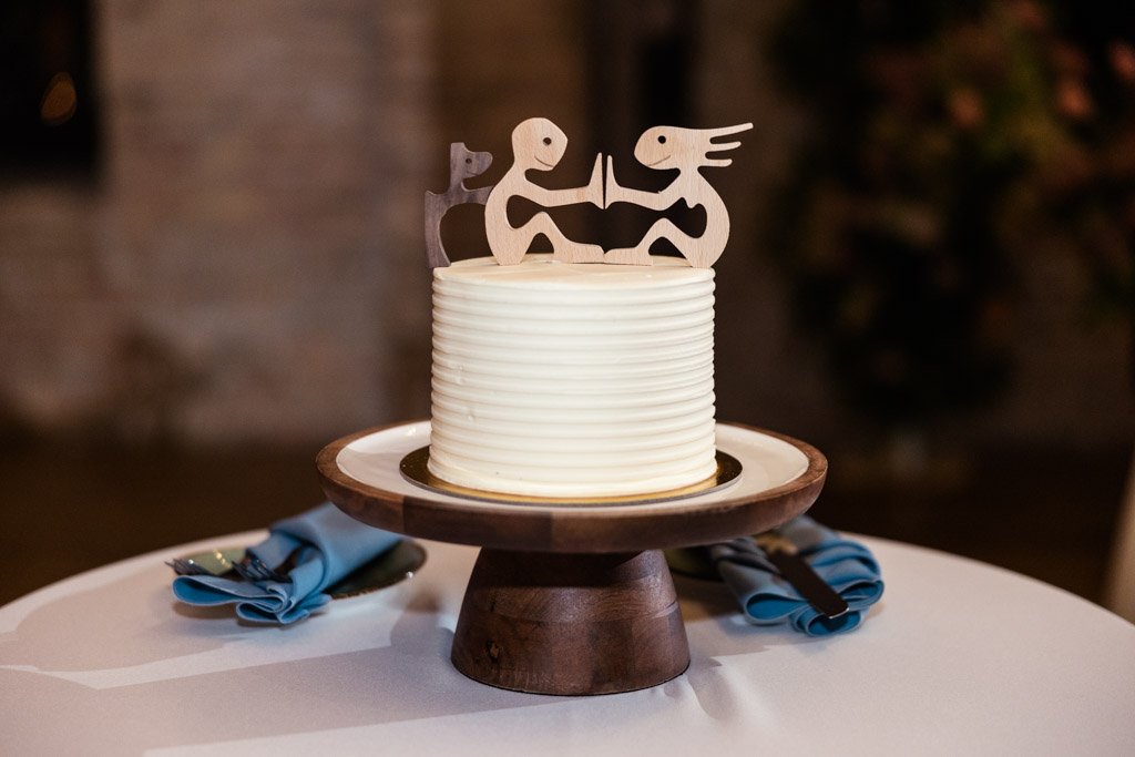 White wedding cake with ridged frosting, topped by custom wooden cake topper and set on a wooden stand for Lacuna Lofts wedding reception