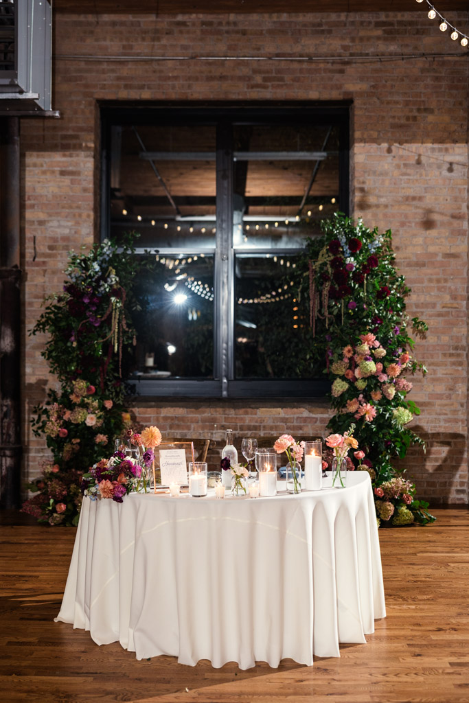 Sweetheart table with white tablecloth, floral arrangements, and candles, set in front of a window and brick wall in Lacuna Lofts wedding reception space