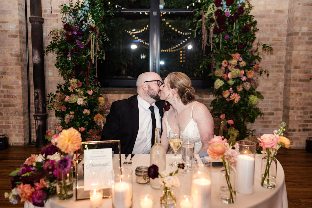 Bride and groom kiss at a decorated sweetheart table with flowers and candles during their wedding reception at Lacuna Lofts