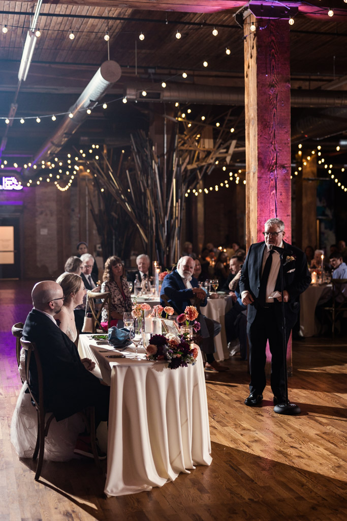 Father of the bride gives a speech at a Lacuna Lofts wedding reception with guests seated at decorated tables under string lights