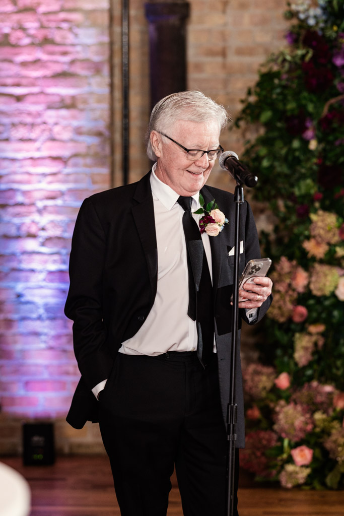 Older man in a suit reads from his phone at a microphone, smiling, with floral decor and a brick background at Lacuna Lofts.