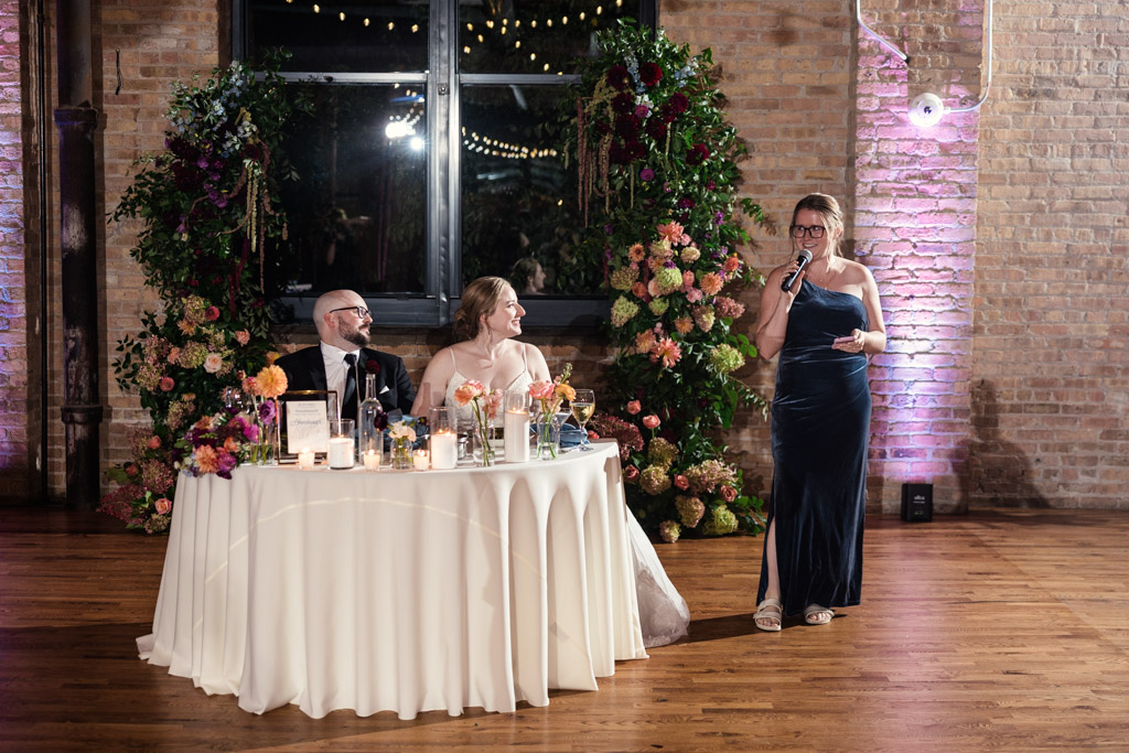 Maid of Honor in a navy dress gives a speech while bride and groom listen during their wedding reception at Lacuna Lofts