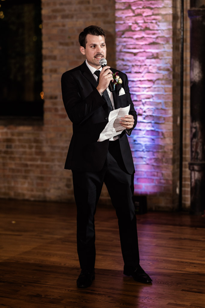 Best Man in a suit holding a microphone and speech notes, speaking indoors at Lacuna Lofts against a brick wall with colored lighting