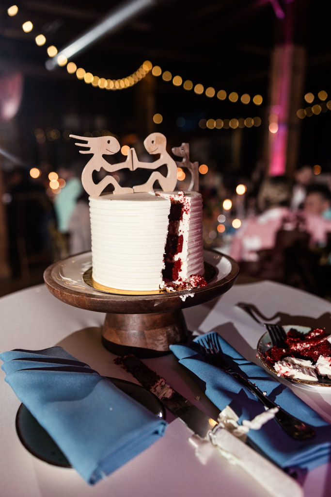 White wedding cake with a slice missing, custom wooden cake topper, blue napkins, and string lights in the background during Lacuna Lofts wedding reception