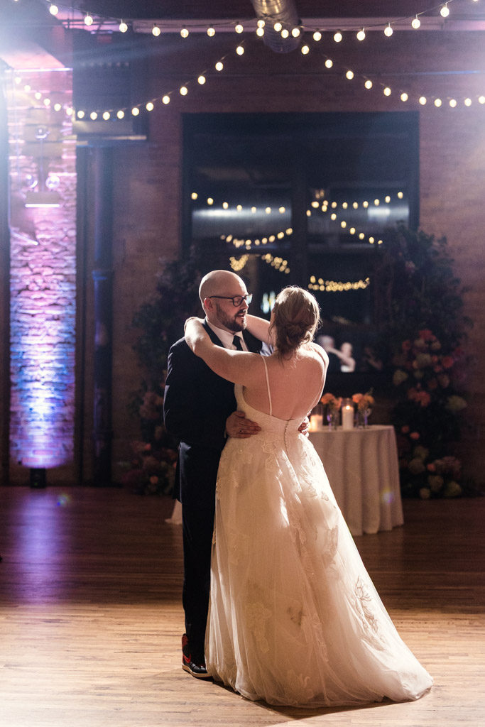 Bride and groom share their first dance under string lights in the warmly lit wedding reception space at Lacuna Lofts