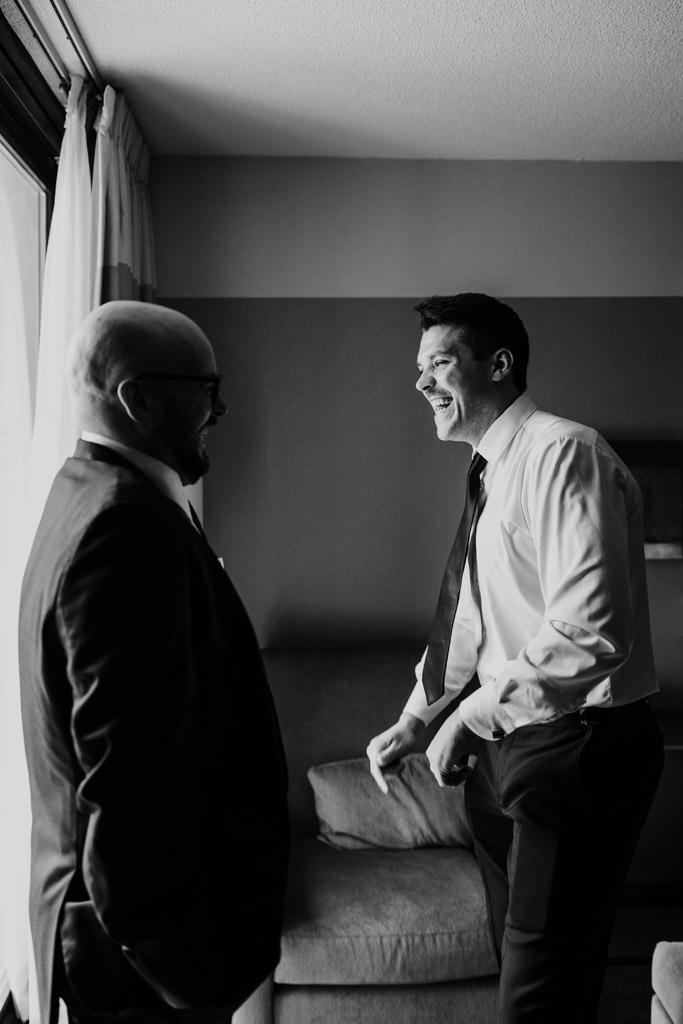 Black and white photo of groom and Best Man in formalwear laughing by a window in a room at Crowne Plaza West Loop in Chicago