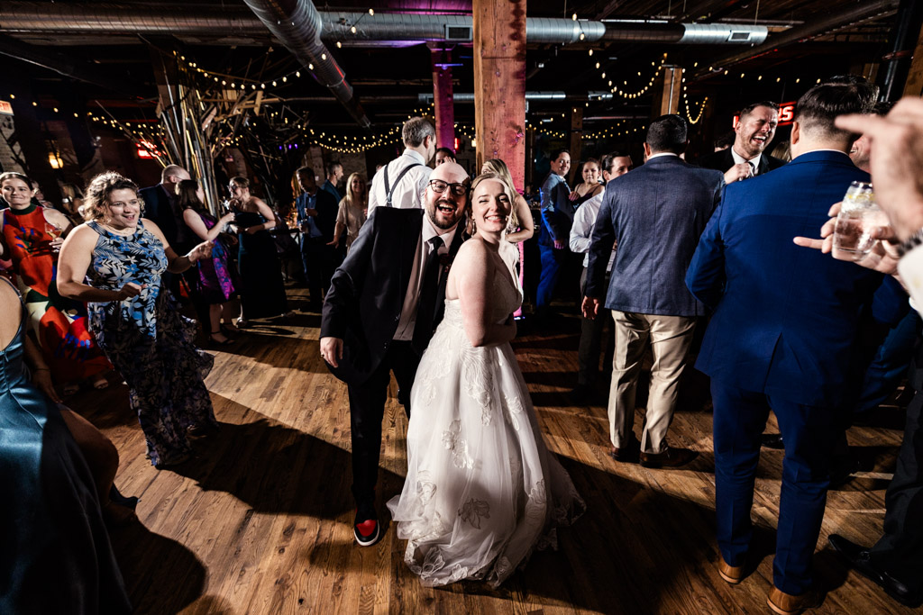 Bride and groom smile on a lively Lacuna Lofts dance floor surrounded by wedding guests in formal attire