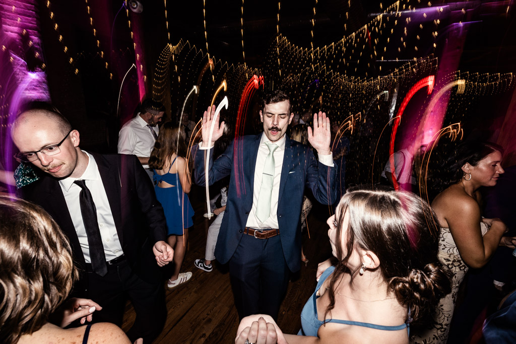 Guests in formal attire dance under string lights at Lacuna Lofts, with colorful light streaks and blurred motion effects