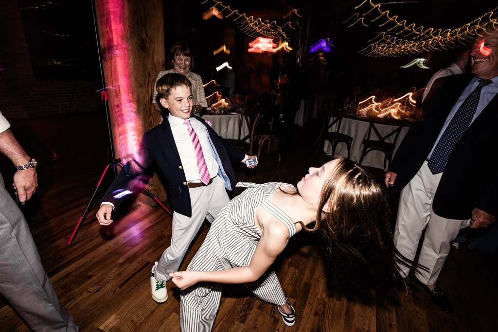 Two kids energetically dance together at wedding reception in Lacuna Lofts, surrounded by laughing adults and colorful lights