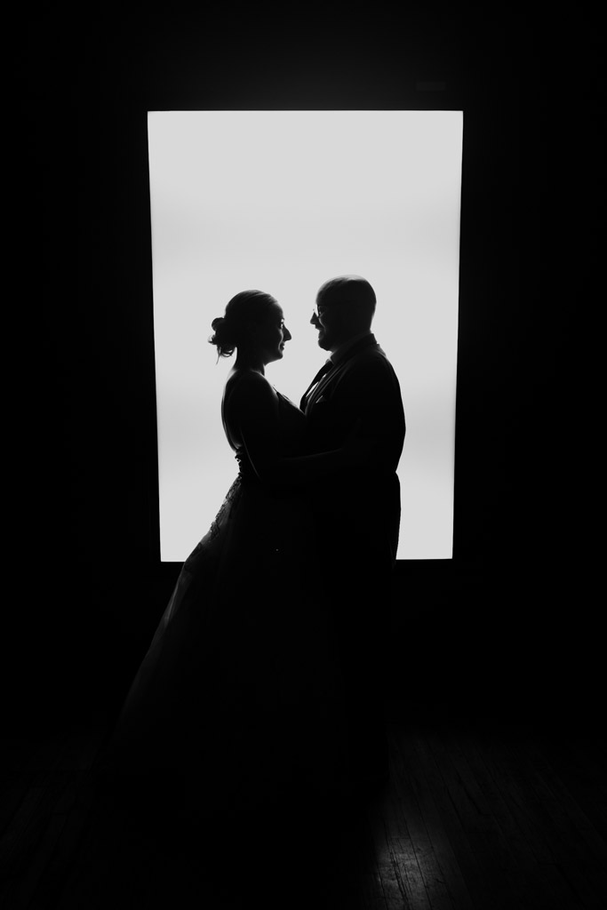 Silhouetted newlywed couple faces each other in front of a bright rectangular light in a dark room at Lacuna Lofts