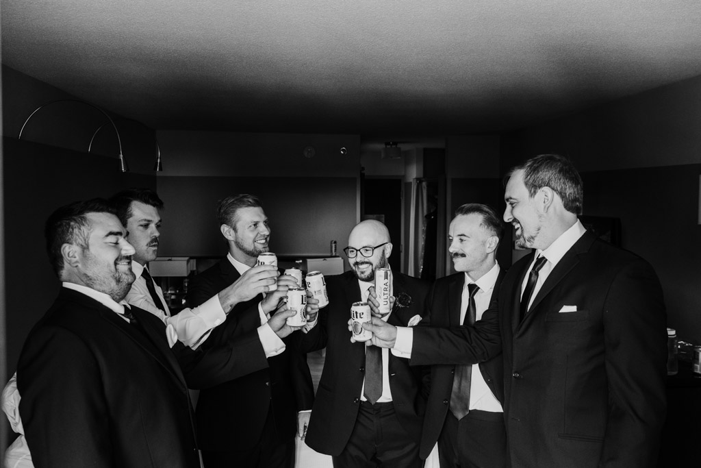 Black and white photo of groom and groomsmen in suits smile and toast with cans of beers in a dimly lit room at Crowne Plaza West Loop before wedding celebration at Lacuna Lofts