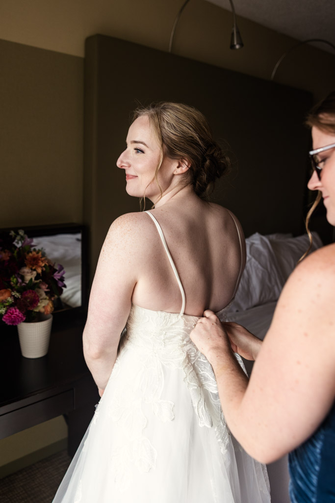 Bride smiles as Maid of Honor helps button the back of her white wedding dress while getting ready at Crowne Plaza West Loop before her wedding celebration at Lacuna Lofts