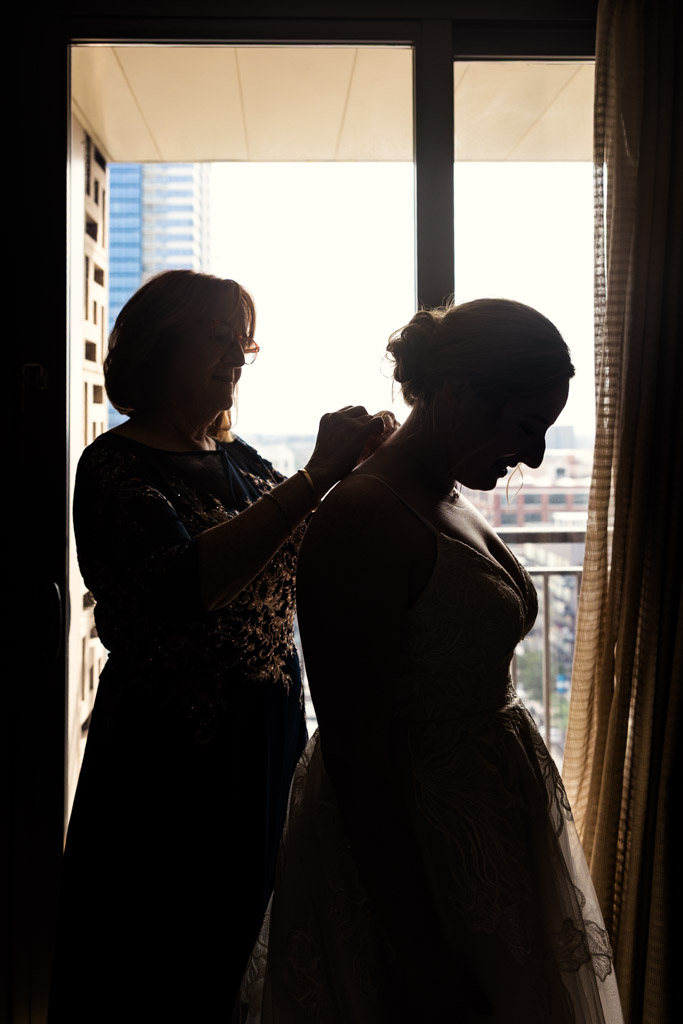 Creative silhouette photo of bride's mother helping bride with her dress near a bright window while getting ready at Crowne Plaza West Loop before Lacuna Lofts wedding celebration