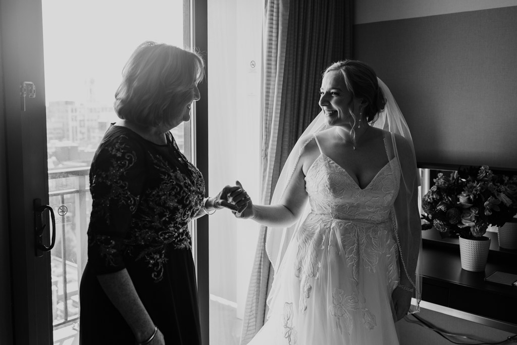 Black and white photo of bride in a wedding dress holds hands and smiles at a woman by a sunlit window at Crowne Plaza West Loop while getting ready for her Lacuna Lofts wedding celebration