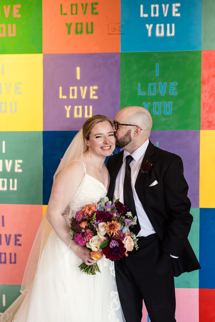 Groom kisses bride's cheek while they stand in front of a colorful 
