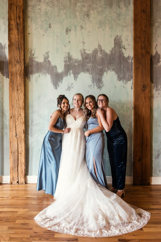 Bride and bridesmaids in formal dresses smile and pose together indoors at Lacuna Lofts, set against a rustic, textured wall and wooden beams