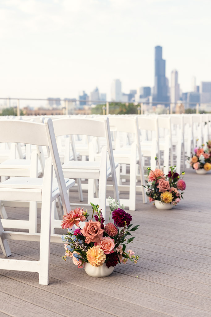 White chairs with flower arrangements in rooftop wedding ceremony space at Lacuna Lofts with the Chicago skyline visible in the background