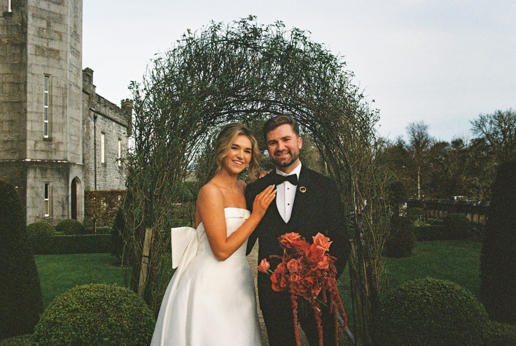 Bride and groom pose, smiling, in front of a floral arch in a garden near the stone walls of Markree Castle