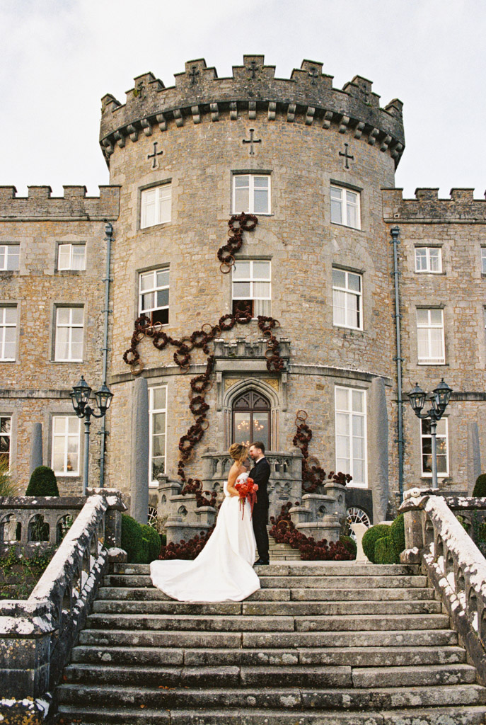 A bride and groom stand on the steps of Markree Castle, embracing in front of the grand stone facade adorned with floral decor