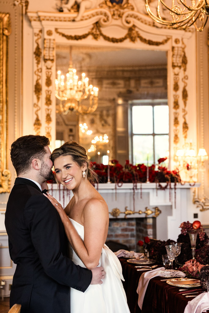 A bride and groom embrace, smiling, in an ornate, elegant wedding reception space with chandeliers and a decorated table at Markree Castle