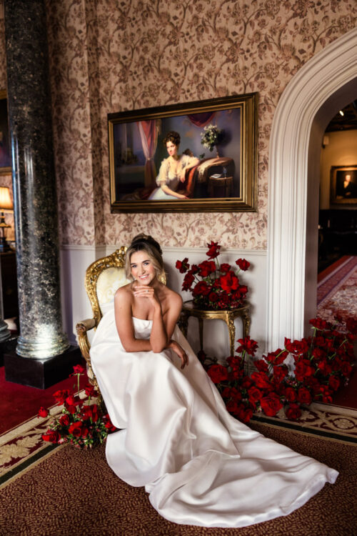 Bride in a white gown sits on an ornate chair, surrounded by red roses, in an elegant, vintage room—capturing the timeless charm of a Markree Castle wedding.